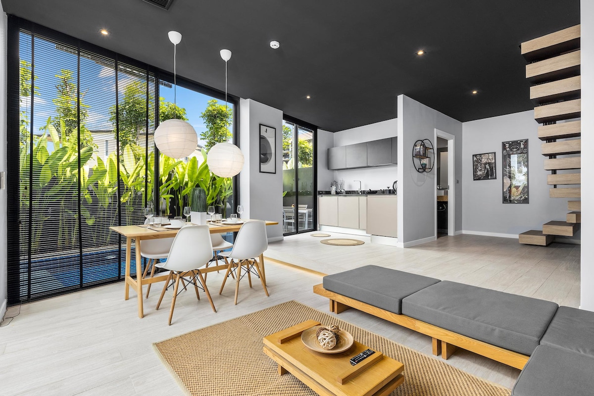 A spacious living area features a minimalist design with natural light flooding in through large glass doors. A simple dining table and chairs are positioned near a stylish sofa. Lush greenery is visible outside, enhancing the serene atmosphere.