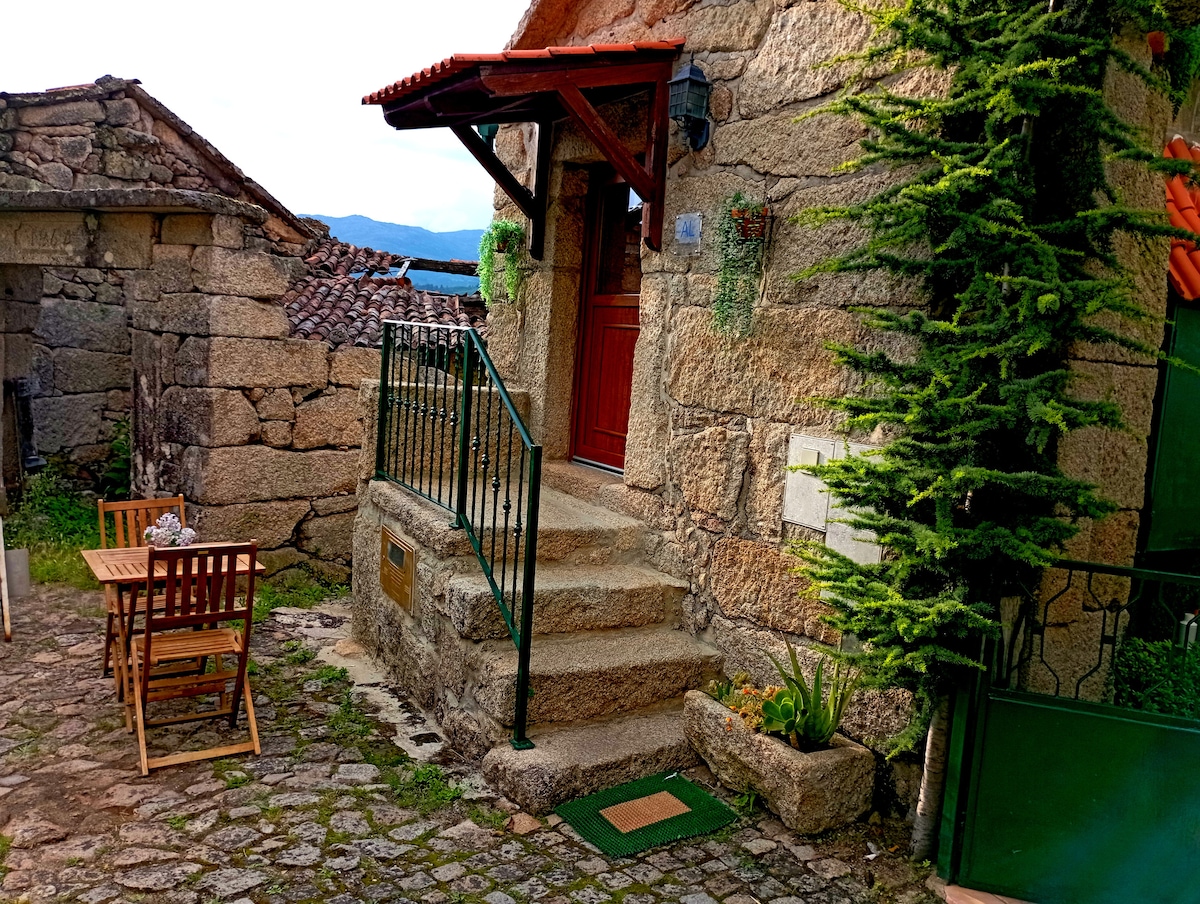 A stone entrance is framed by a red wooden door and a small set of steps leading to the home. To the side, a wooden table and chairs are placed on a cobblestone patio, complemented by greenery and rustic stone walls.