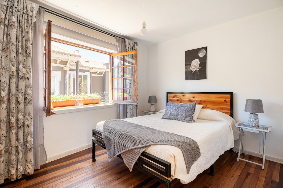A bright bedroom features a spacious bed with a wooden headboard. Natural light filters in through a window adorned with floral curtains, which are opened to reveal an inviting view. A small bedside table with a lamp is positioned on each side of the bed.