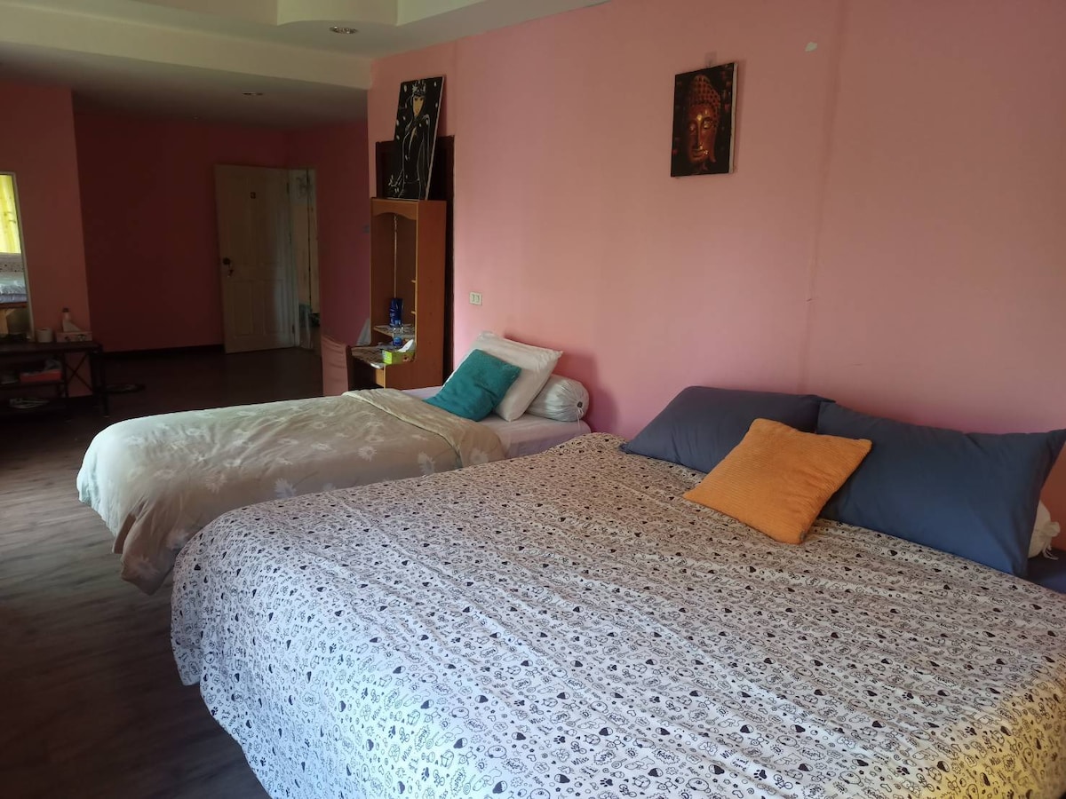 A guest room features two beds, one large with a decorative blanket and one smaller bed with a light-colored cover. Bright pink walls create a vibrant atmosphere, complemented by decorative artwork. A window allows natural light to enter, enhancing the cozy feel of the space.