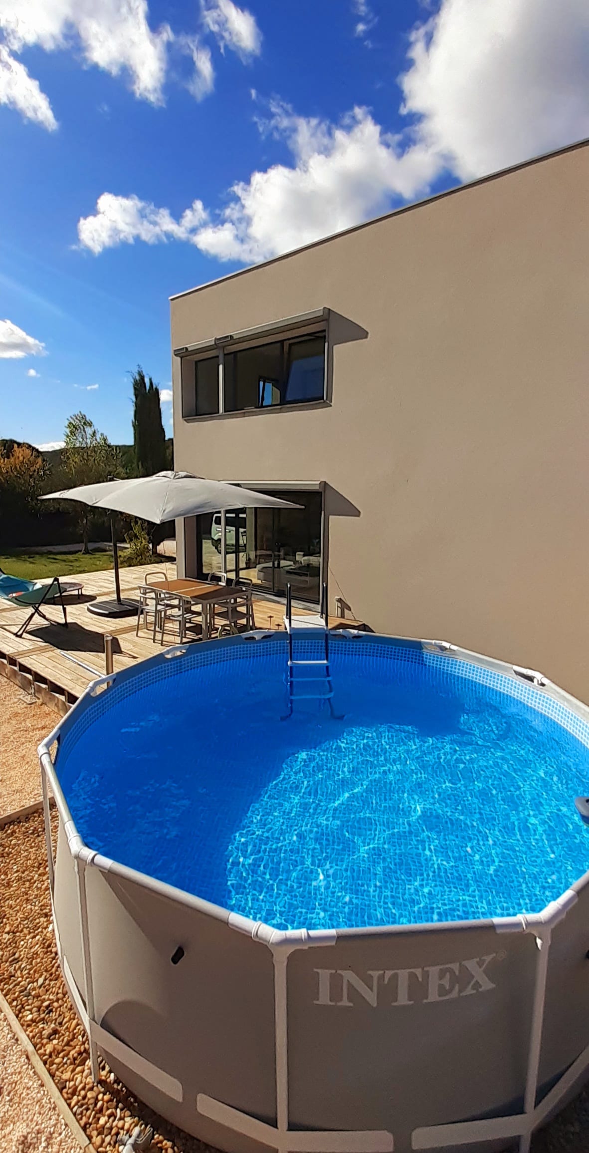 A circular, above-ground pool is surrounded by a wooden deck, inviting relaxation. Sunlight reflects off the clear water, while a shaded seating area is visible nearby. Large glass doors lead into the modern home, enhancing the outdoor living experience.