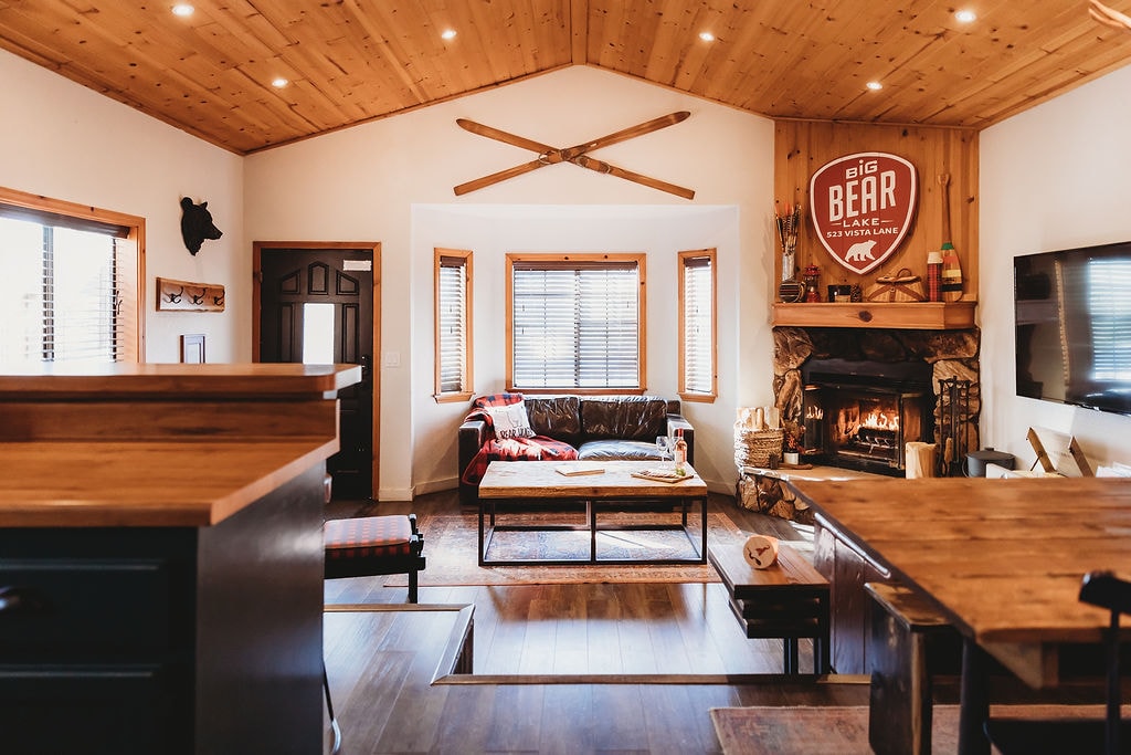 An inviting open concept living area is showcased, featuring a large native stone fireplace and a comfortable leather couch. Vaulted pine ceilings highlight the spacious layout, while light streams in through large windows, enhancing the cozy atmosphere.
