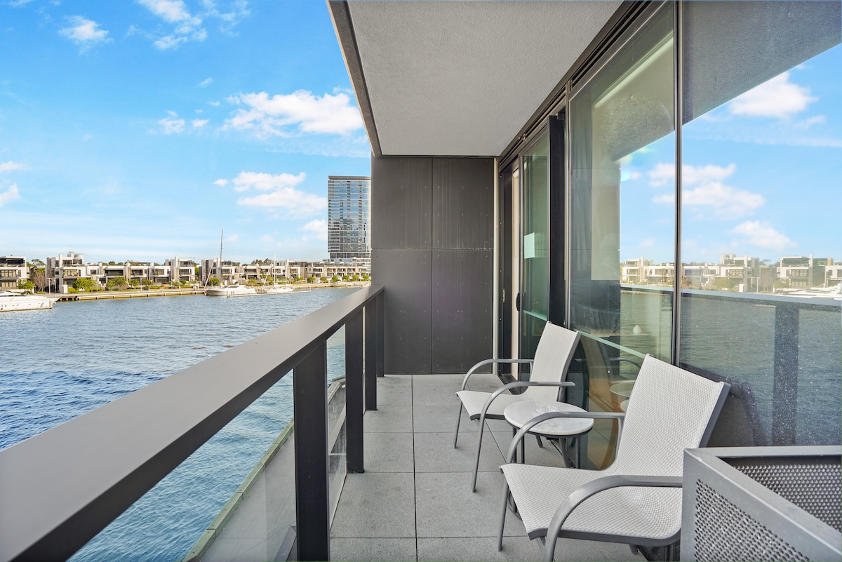 A spacious balcony is showcased, featuring two white chairs and a small table. The waterfront view is highlighted, with blue water and distant buildings visible across the river. Large glass doors connect the balcony to the interior, allowing natural light to flow in.