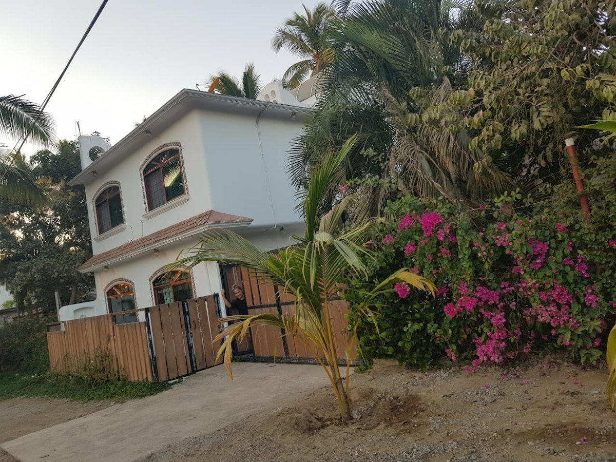 A two-story house is surrounded by lush greenery and vibrant flowering plants. The home's exterior features large windows that allow natural light to enter. A wooden fence provides privacy, and a pathway leads to the entrance, contributing to the inviting atmosphere of the property.