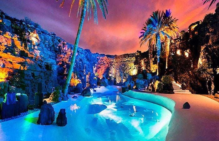 A dramatic landscape features a swimming pool surrounded by natural rock formations and palm trees. The scene is illuminated with vibrant colored lights reflecting off the water, creating a serene and inviting ambiance against a twilight sky.