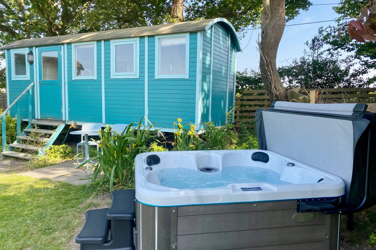 An extra-large shepherd's hut painted in a soothing turquoise sits nestled among greenery. A private hot tub with bubbling water is positioned nearby, providing a tranquil space for relaxation. The scene is framed by trees and a clear blue sky.