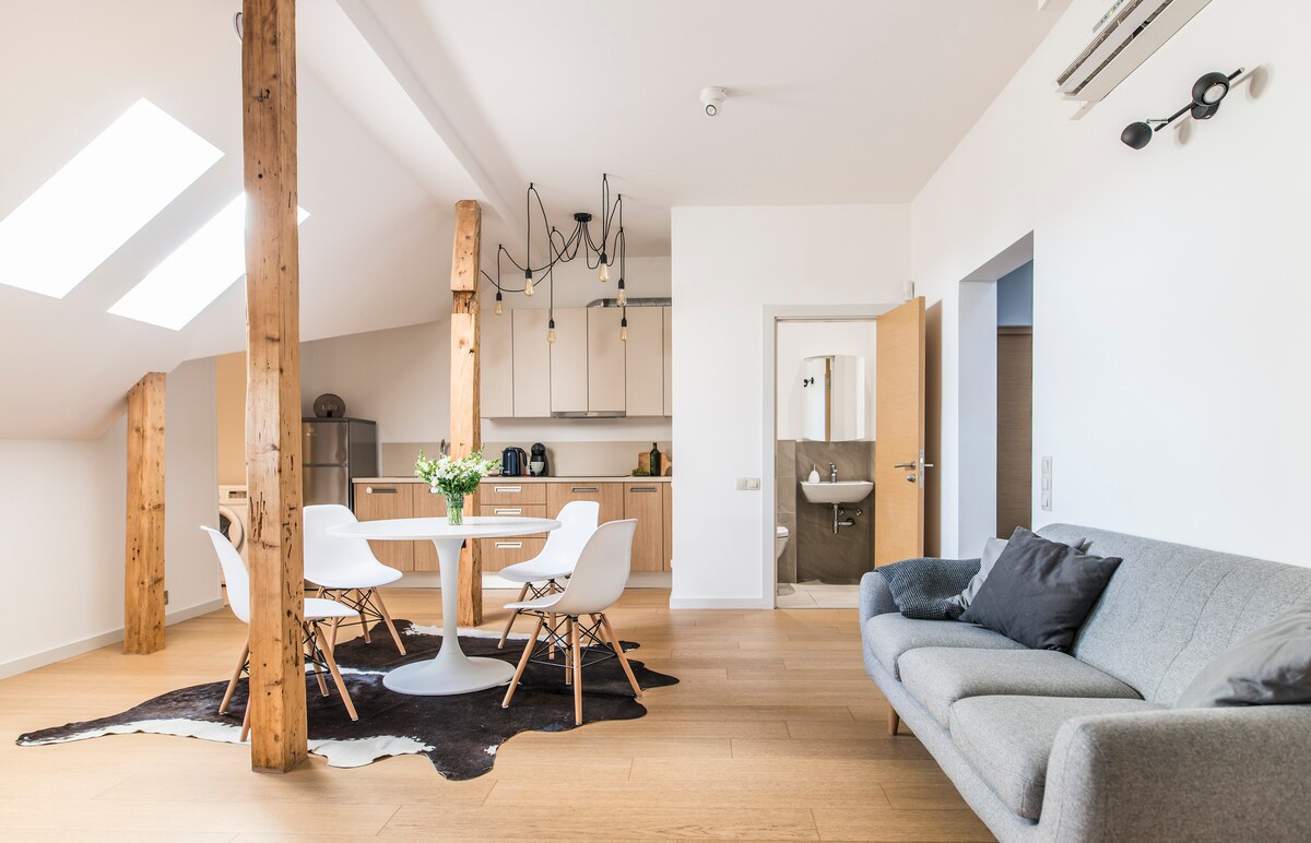A bright living space is furnished with a round dining table surrounded by white chairs, complemented by a gray sofa. Skylights above provide natural light. The open kitchen area is visible in the background, along with a view of a bathroom door.