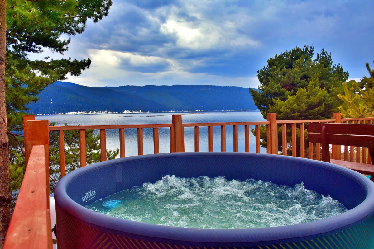 A hot tub with bubbling water is positioned on a deck overlooking a serene lake surrounded by hills. The natural landscape is highlighted by green trees and distant mountains under a cloudy sky.