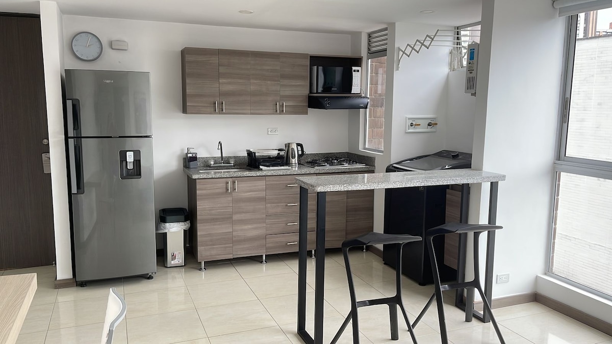 A modern kitchen setup features sleek cabinets and a granite countertop. Stainless steel appliances, including a refrigerator and a stove, are visible. A small bar with two stools provides additional seating. Natural light streams through large windows, enhancing the spacious feel of the area.