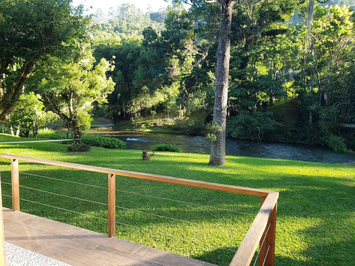 A view from a wooden deck overlooking a lush green yard and a gently flowing river bordered by trees. The vibrant grass and distant foliage create a serene natural setting.