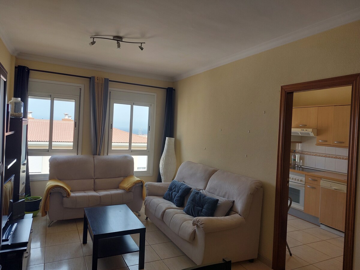 A bright living area features two comfortable sofas positioned around a black coffee table. Natural light enters through large windows, revealing views of rooftops. A doorway leads to a kitchen area, while a simple floor lamp adds to the inviting atmosphere.