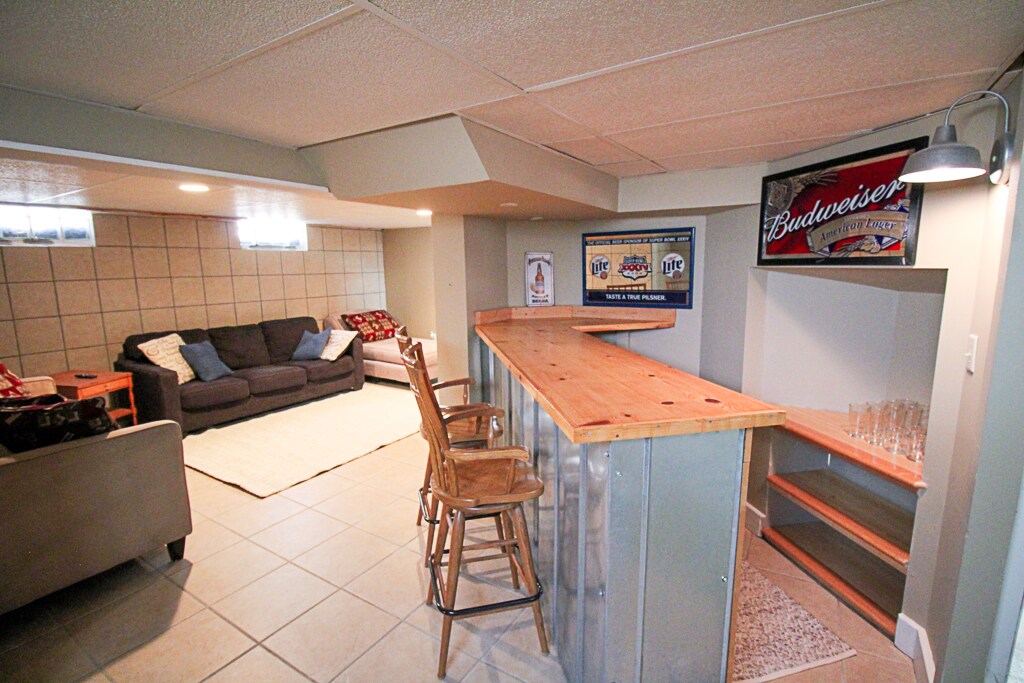 A spacious basement area features a comfortable seating arrangement with a dark sofa and a light rug. A wooden bar with metal accents is positioned against one wall, accompanied by three high stools. Wall-mounted art adds character to the space, and natural light enters through small windows.