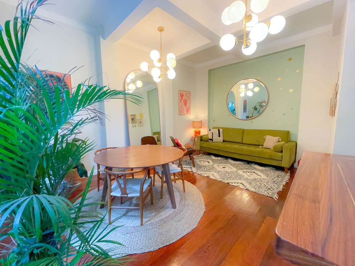 A brightly lit living area features a cozy green sofa and a round wooden dining table surrounded by chairs. Plants embellish the space, and a large round mirror on the wall reflects the elegant light fixtures. A textured area rug adds warmth to the polished wooden floor.