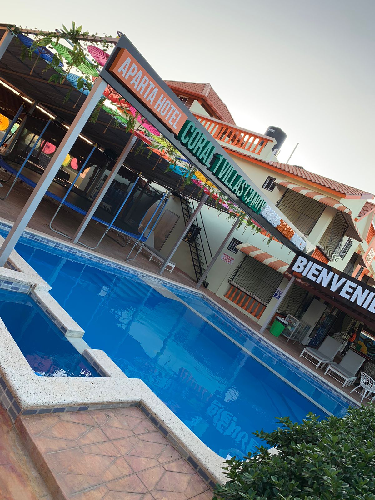 A welcoming entrance to the hotel is shown, featuring a colorful sign that reads 'Bienvenido.' A refreshing pool is surrounded by lounge chairs and shaded seating areas, while the two-story building provides ample outdoor space for relaxation.