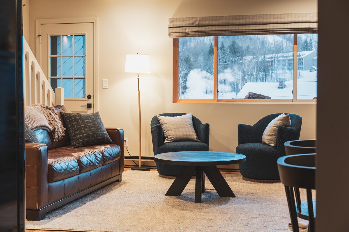 A living area features a leather sofa and two accent chairs, complemented by a circular coffee table. Natural light floods the space through a large window with mountain views. A floor lamp provides additional lighting, and a textured rug adds warmth to the room.