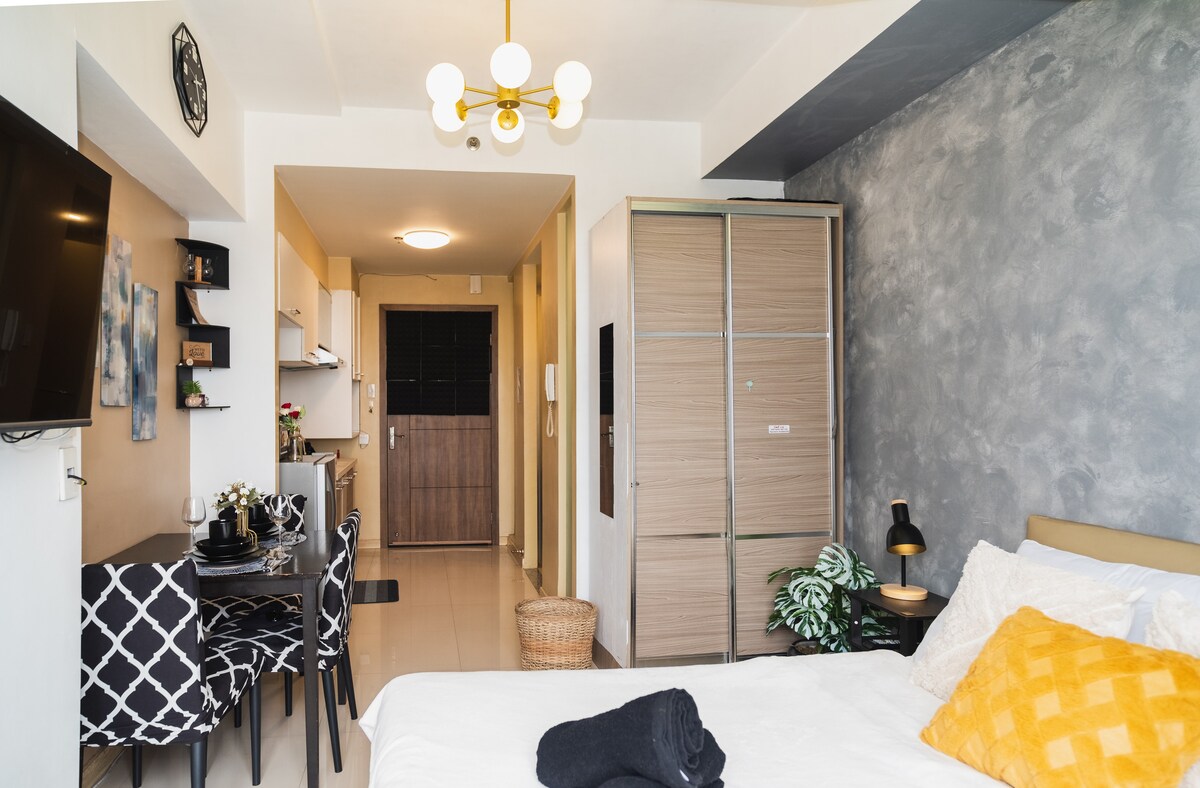 An open-plan studio is shown, with a comfortable bed dressed in white linens and decorative pillows. A small dining area with a round table and chairs is visible, along with a kitchenette equipped with basic appliances. A wall-mounted TV and a closet complete the space.