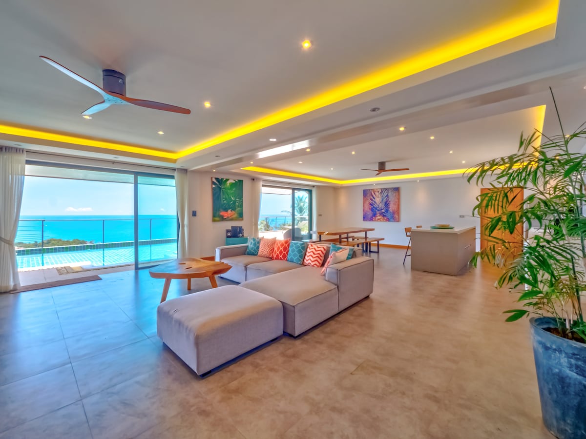 An open living space offers panoramic views of the ocean, complemented by a large sofa with colorful cushions. Natural light floods the room through floor-to-ceiling sliding glass doors, while a modern kitchen and dining area are visible in the background.