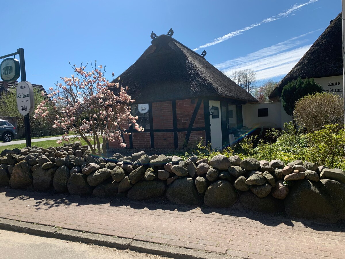 The thatched house is framed by a stone wall and features a quaint entrance marked by a sign. In the foreground, a flowering tree adds a touch of color, while greenery surrounds the property, contributing to a tranquil outdoor setting.