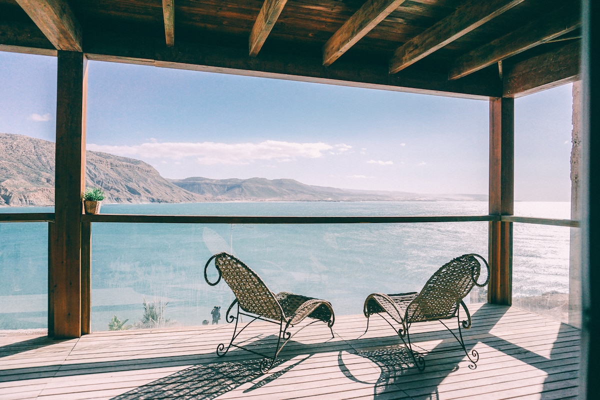 Two woven lounge chairs are positioned on a spacious terrace, offering stunning views of the ocean and surrounding coastline. The seating area is framed by wooden beams, with natural light illuminating the deck. Greenery is visible on a ledge, complementing the serene surroundings.
