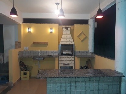 The kitchen area features a granite countertop and a built-in brick barbecue grill. Soft lighting is provided by pendant lamps, and an open window allows natural light to enter. Shelves are mounted on the wall for additional storage.
