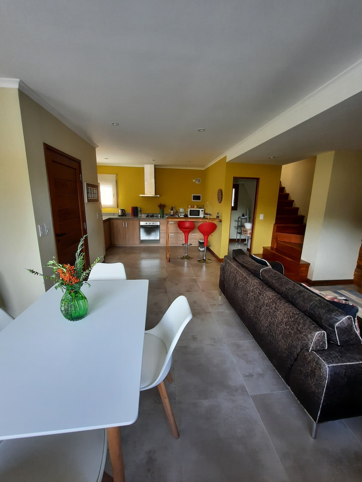 A modern open-concept living area features a dining table with white chairs and a green vase of flowers, connected to the kitchen with wooden cabinetry and modern appliances. Two red bar stools add a pop of color near the kitchen island, while a staircase leads to the upper level.