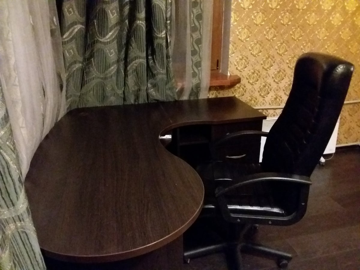A work desk with a curved design is displayed, accompanied by a black ergonomic chair. The backdrop features patterned curtains, and the wall is adorned with subtle golden wallpaper, providing a balanced contrast to the dark wooden tones of the furniture.