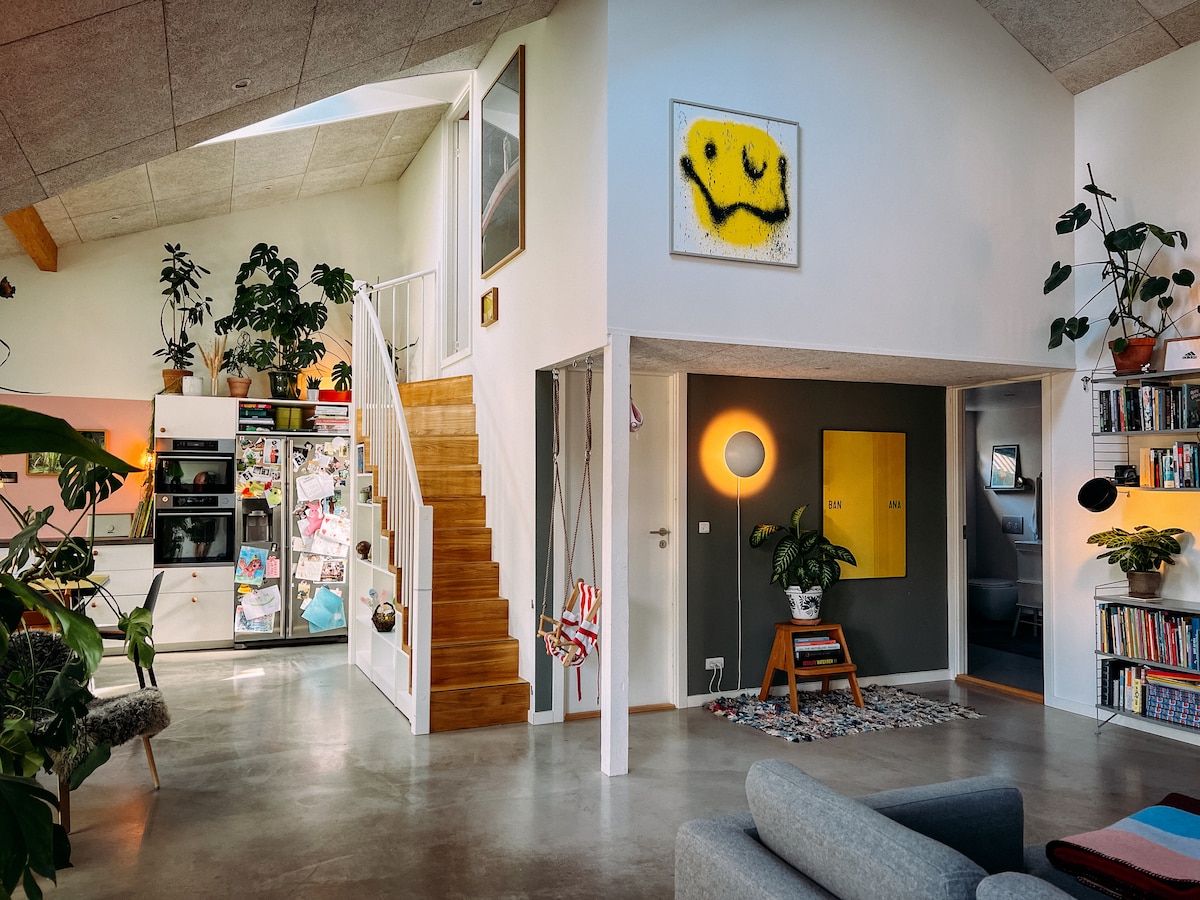 An open living area is visible, featuring high ceilings and numerous plants. A colorful artwork hangs on the wall above a sleek console table. A comfortable sofa faces the kitchen, which includes modern appliances and bright cabinets. Stairs lead to the upper level, enhancing the spacious feel.