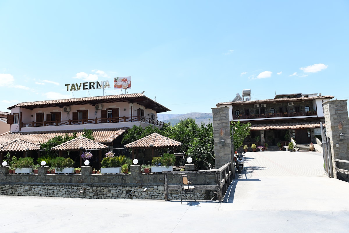 The image depicts a two-story taverna building, featuring a stone facade and wooden balconies. A covered outdoor seating area with planters filled with flowers is visible in the foreground. A pathway leads to the entrance, where additional seating and decorative elements can be seen.
