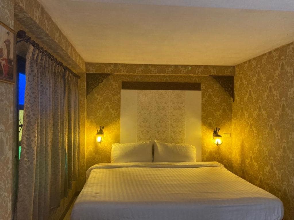 A sleeping area is presented with a spacious bed, framed by elegant patterned wallpaper and soft lighting from wall-mounted lamps. A large window with sheer curtains allows for natural light, while a decorative tapestry adds a touch of charm.