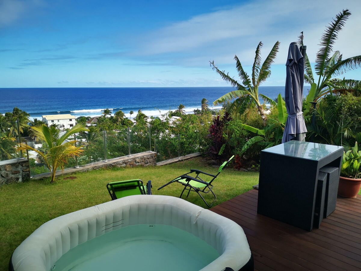 A private outdoor space is presented, featuring a traditional hot tub and a dining area with a table and chairs. Verdant tropical plants surround the area, and the vast ocean is visible in the background, extending to the horizon under a clear blue sky.