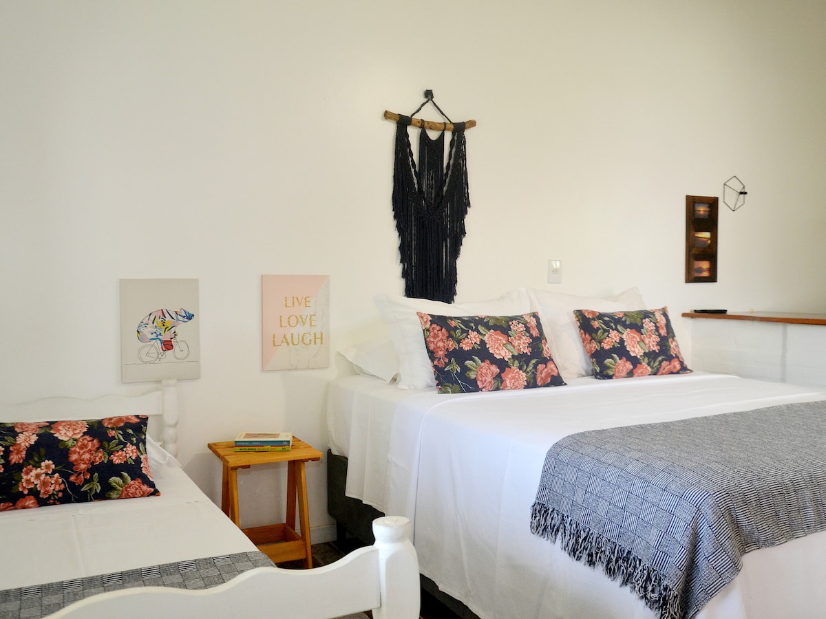 An integrated sleeping area features a spacious double bed and a single bed, adorned with floral-patterned pillows. A minimalistic wall art piece and decorative fabric hang above the bed, contributing to the bohemian style. A nightstand is positioned beside the single bed.