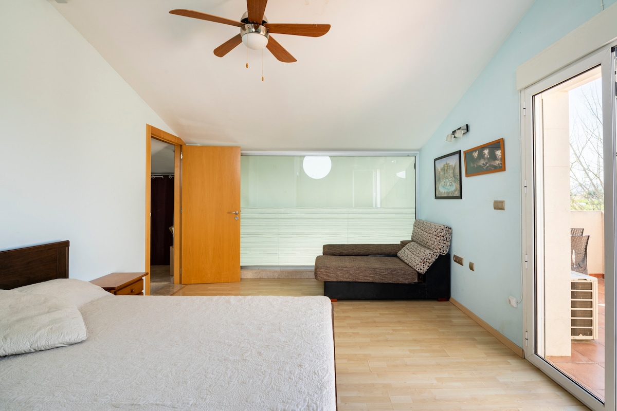 A spacious bedroom features a comfortable bed and a lounging area with a couch. Natural light illuminates the room through large glass doors leading to a private balcony. A ceiling fan adds to the airy feel, while wooden and glass elements create a serene ambiance.