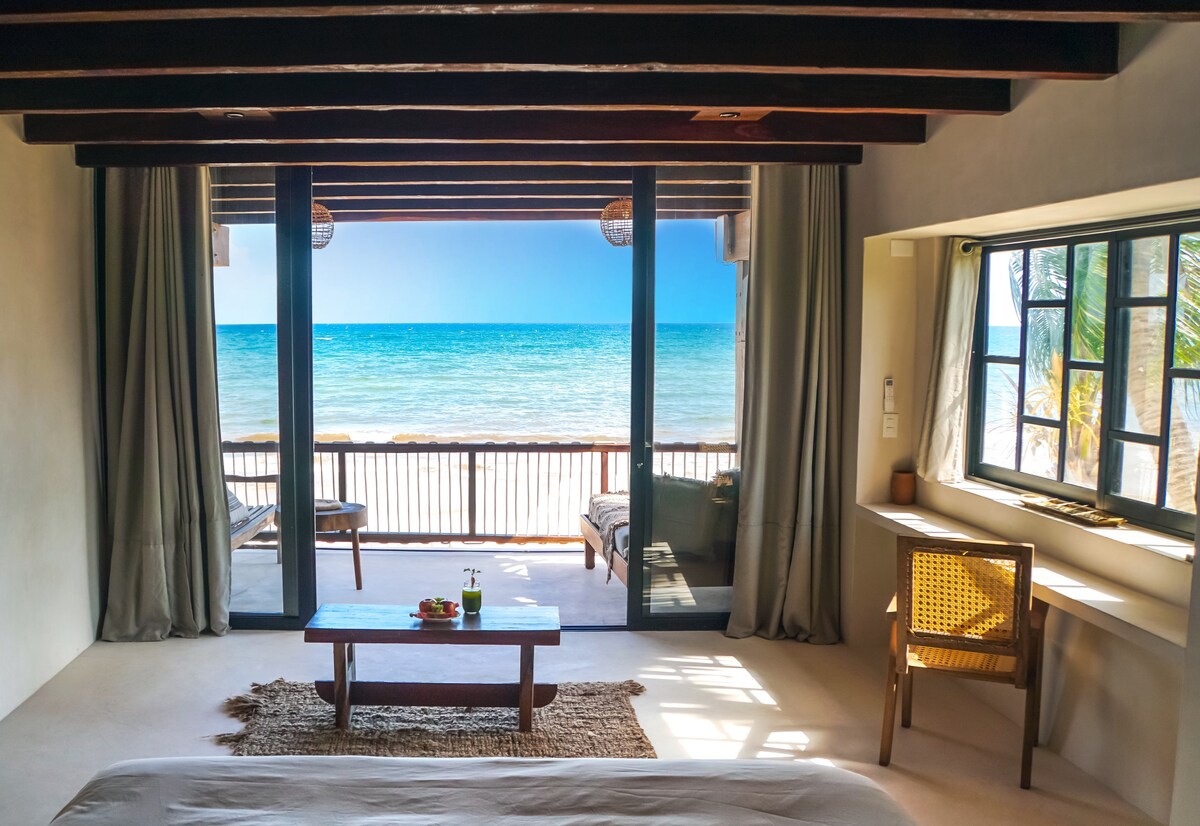 A spacious room features large glass doors opening to a private terrace overlooking the sea. Earthy tones are used throughout, with a cozy bed and a small table positioned in front of the view. Natural light fills the space, enhancing the connection with the outdoor scenery.