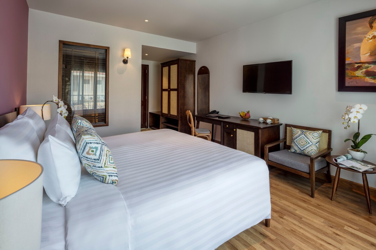 The room features a spacious bed with crisp white linens and decorative pillows. A wall-mounted television is positioned above a wooden desk and chair. Natural light enters through a window, while light wood flooring adds warmth to the space. Art pieces and plants enhance the decor.