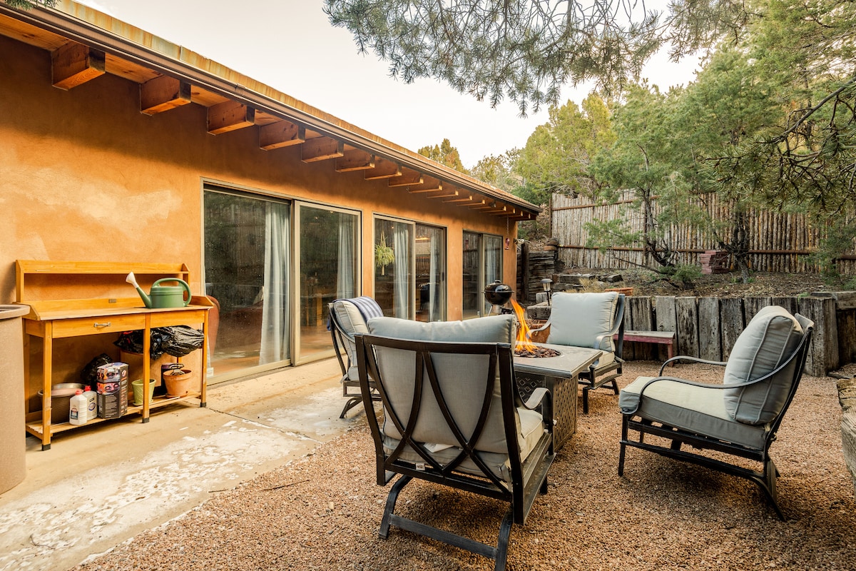 An outdoor patio area features comfortable seating arranged around a fire pit, inviting relaxation. The adobe home is visible in the background, showcasing large sliding doors that open to the interior. Natural surroundings enhance the peaceful ambiance, with trees providing shade.