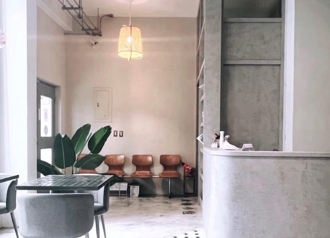 A modern reception area is displayed, featuring a curved counter and a light fixture overhead. Three wooden chairs are arranged against the wall, and a potted plant adds a touch of greenery. Neutral tones create a calm and welcoming environment.