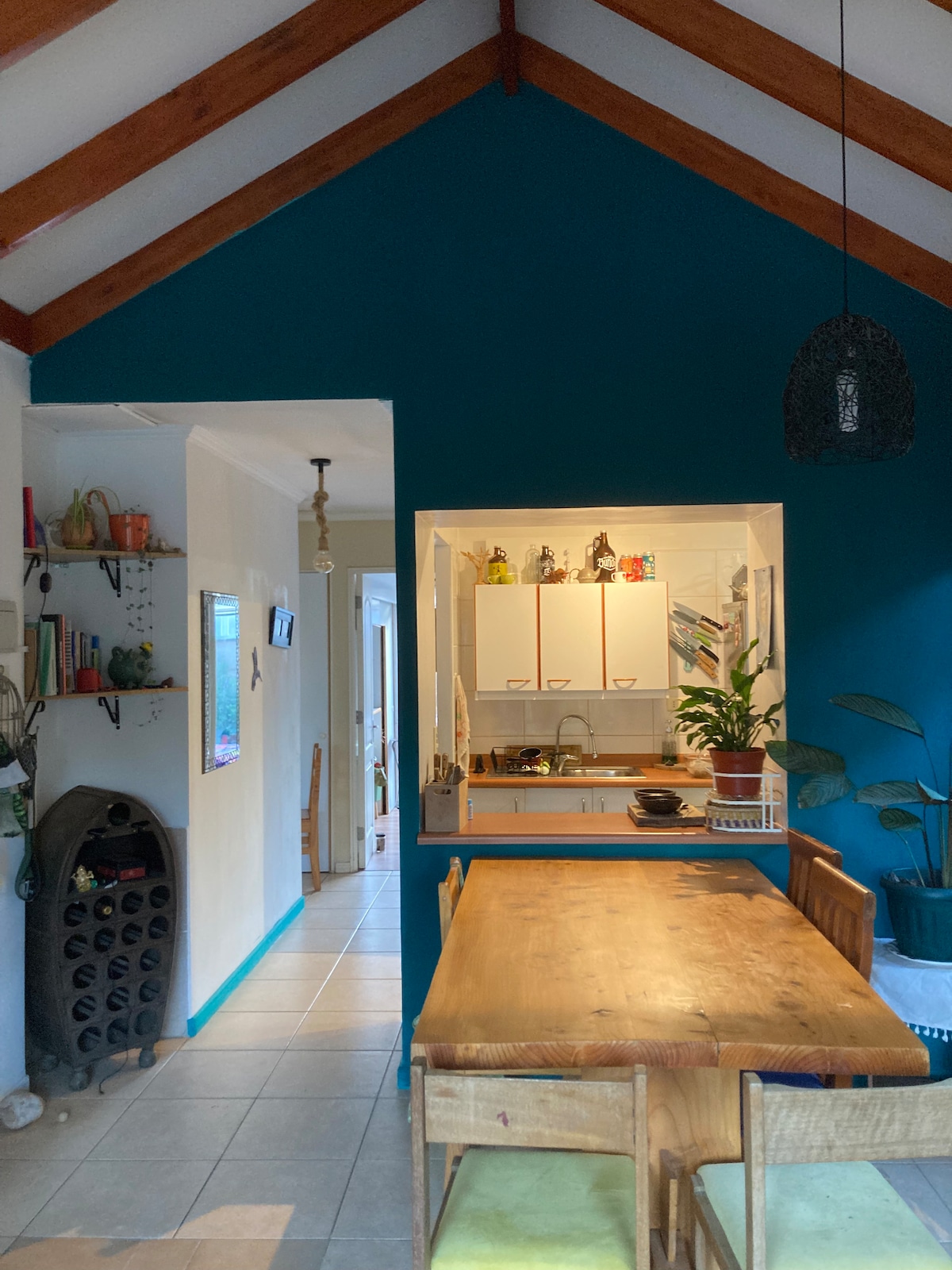 A dining area featuring a wooden table surrounded by chairs offers a gathering space. The walls are painted in a vibrant blue, complemented by wood beams overhead. A glimpse of the kitchen is visible, showcasing white cabinetry and decorative elements. Houseplants add natural touches to the space.