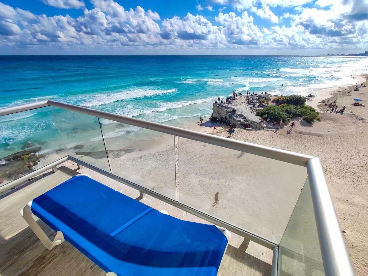 A spacious balcony offers a clear view of the turquoise ocean and sandy beach below. The shoreline is lined with waves, while scattered beachgoers can be seen enjoying the sun. A single blue lounge chair is positioned for relaxation.