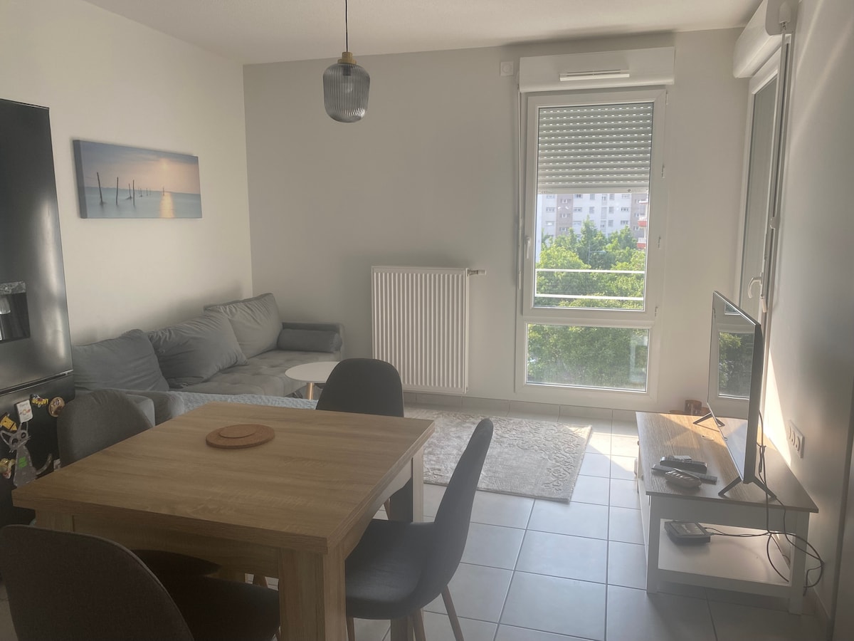 A contemporary living area features a comfortable gray sofa and a light wood dining table with four chairs. Natural light floods the space through a large window that offers a view of greenery. A flat-screen television sits on a minimalist white media console.