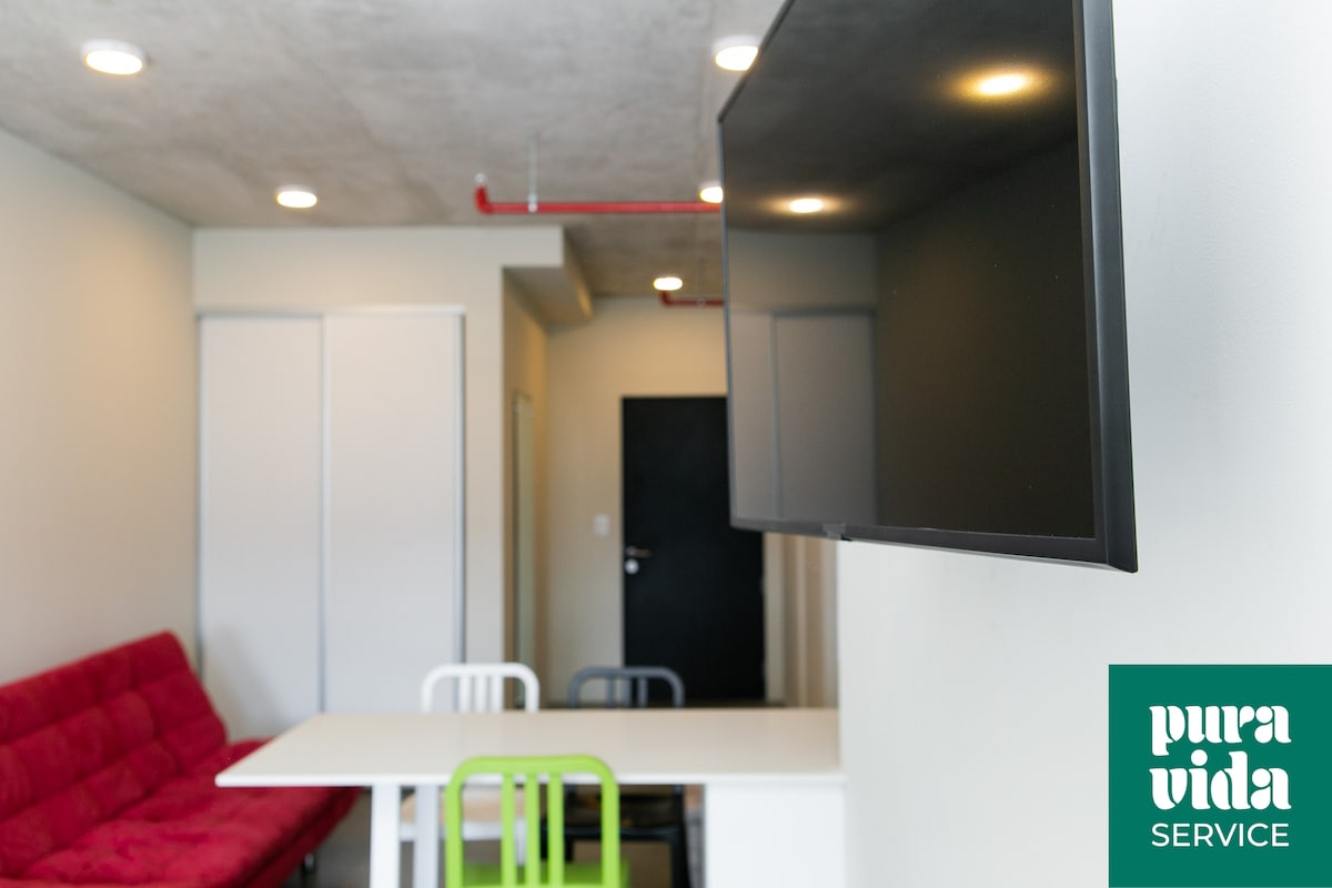A modern living space features a large flat-screen TV mounted on the wall. A white table with colorful chairs is positioned in front of a red sofa bed. The minimalist design is highlighted by smooth concrete ceilings and neutral-colored walls.