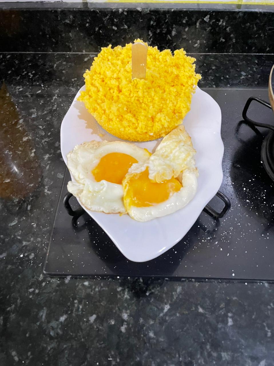 A white dish is shown on a dark countertop, featuring two sunny-side-up eggs with vibrant yellow yolks beside a bowl of yellow rice. The bowl is topped with a wooden stick for serving.