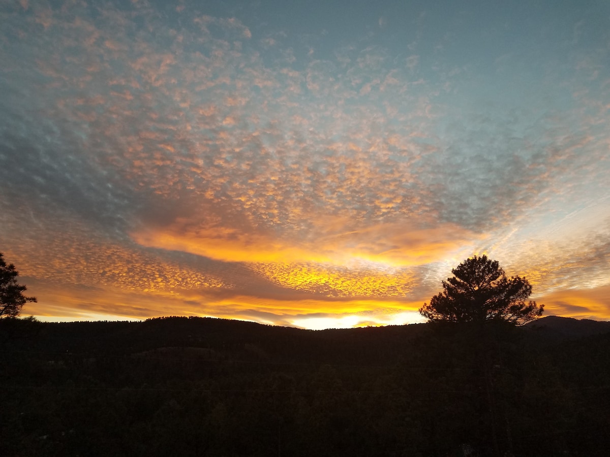 A stunning sunset is captured, showcasing vibrant orange and yellow hues blending into the sky. Fluffy clouds reflect the colors, while silhouettes of surrounding trees and distant mountains provide a natural border to the scene.