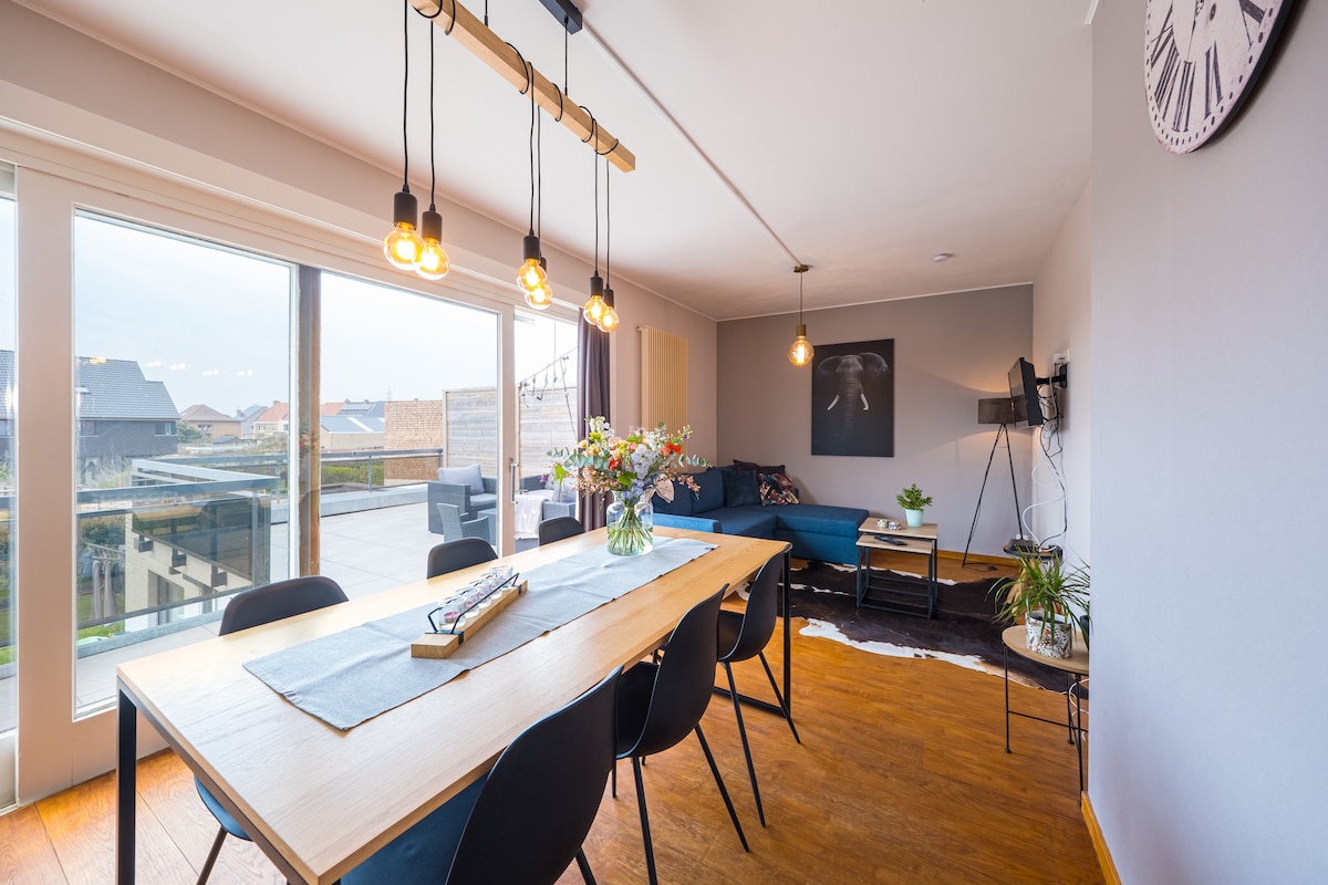 A light-filled living area showcases a long dining table surrounded by black chairs, with a cozy seating arrangement nearby. Large sliding glass doors provide access to a sun terrace, while a decorative wall clock and a floral centerpiece add subtle touches to the space.