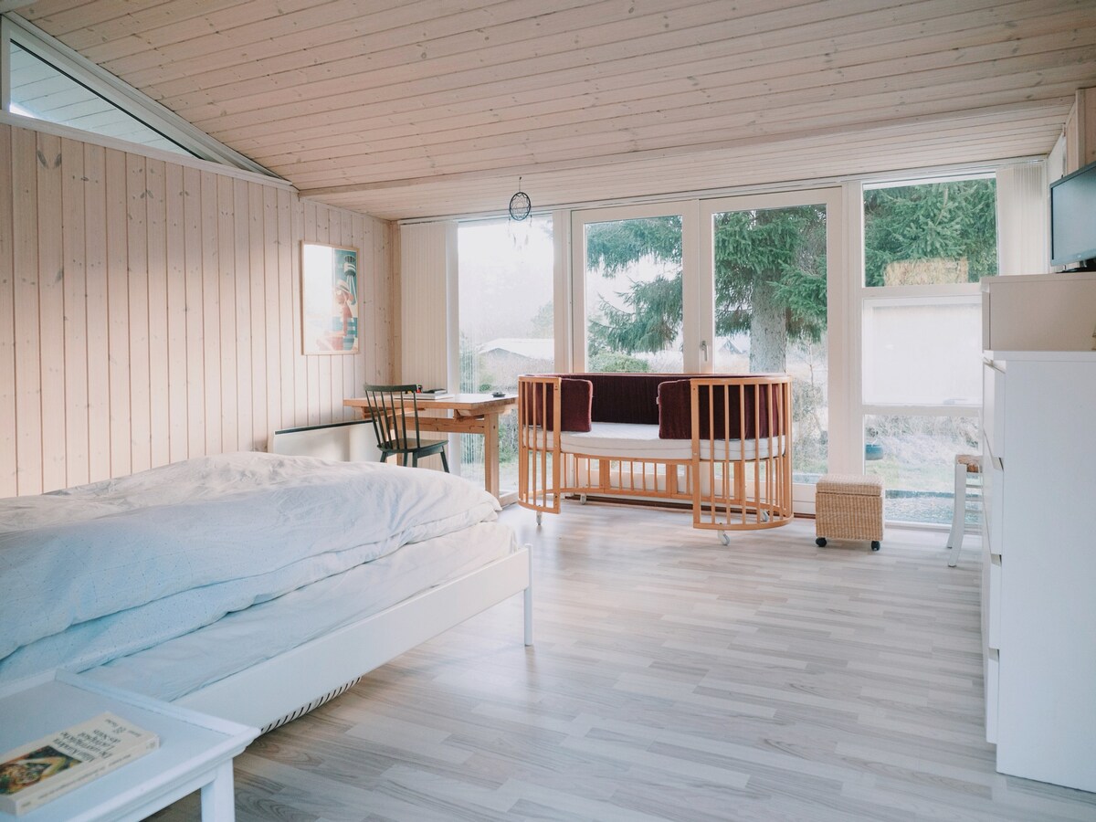 A spacious master bedroom features a large bed with a soft blanket, complemented by a sitting area with a small table and a cozy couch. Large windows allow natural light to fill the room, offering views of the garden outside. Light wood paneling enhances the tranquil atmosphere.