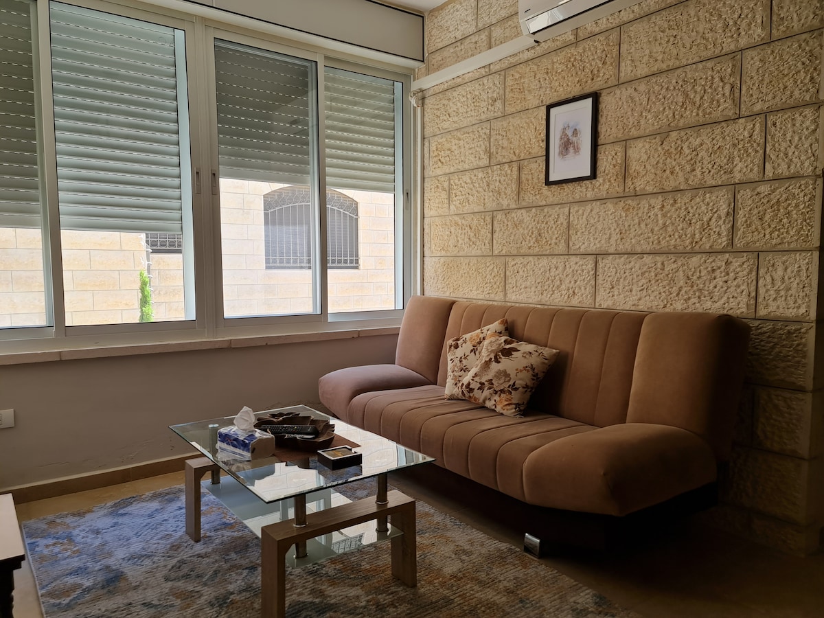 A cozy living area features a brown sofa with decorative cushions, positioned across from a glass coffee table. Natural light floods the space through large windows with silver blinds, complemented by textured stone walls. A framed picture adds a decorative touch to the wall.
