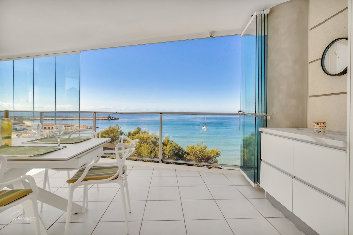 A bright dining area features a white table with four chairs, positioned near expansive glass doors. Stunning views of the Mediterranean Sea are visible through the glass curtains, which can be opened to embrace the outdoors. The space is filled with natural light.