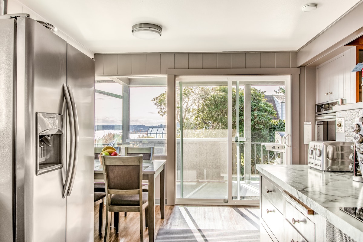A modern kitchen area features stainless steel appliances, including a refrigerator and a microwave. A dining table with four chairs is positioned near sliding glass doors, allowing natural light to fill the space and providing a view of the outdoors.