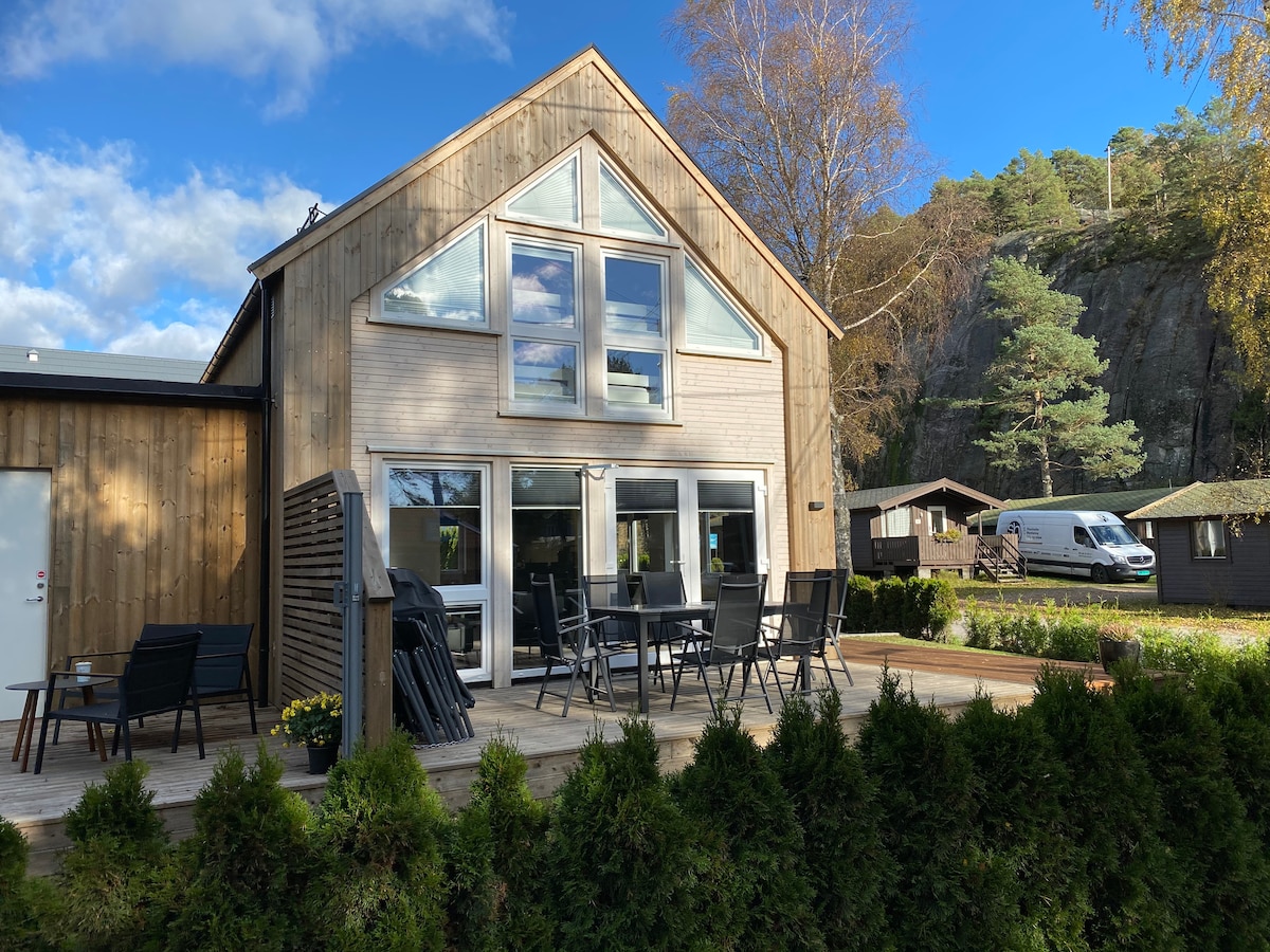 The modern wooden cabin features large triangular windows and a spacious deck, furnished with multiple black chairs and a table. Surrounding greenery adds a natural touch, while neighboring cabins and a vehicle can be seen in the background under a partially cloudy sky.