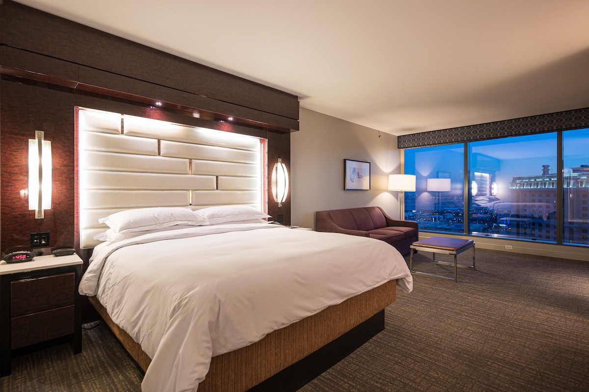 A spacious sleeping area features a king-size bed with crisp linens, complemented by modern bedside lighting. A sleek sofa and coffee table are positioned nearby, with floor-to-ceiling windows offering a view of the Las Vegas skyline, enhancing the overall design of the suite.