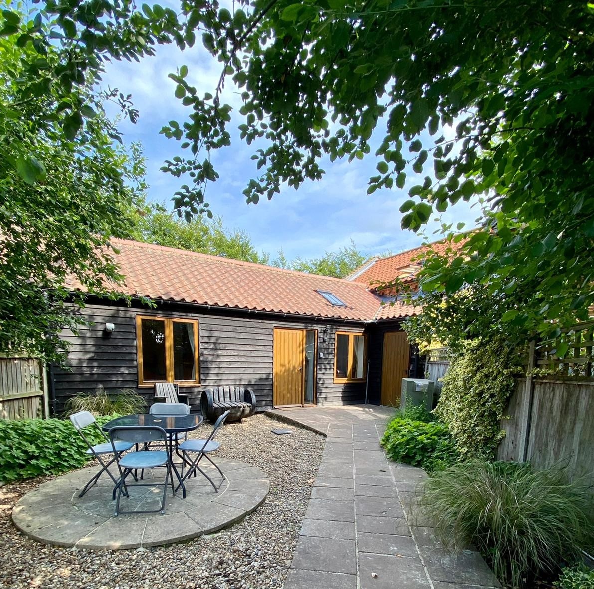 A secluded outdoor area features a gravel pathway leading to a barn-style building. A circular stone patio is surrounded by greenery, with a small table and chairs positioned nearby. Trees provide shade, and wooden doors accent the barn's rustic charm.